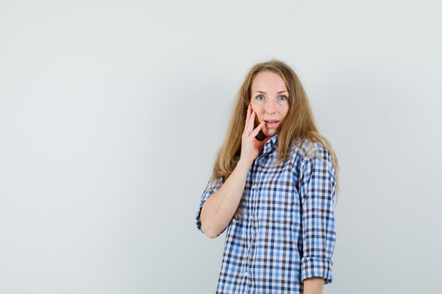 Blonde lady in shirt holding hand on cheek and looking amazed ,