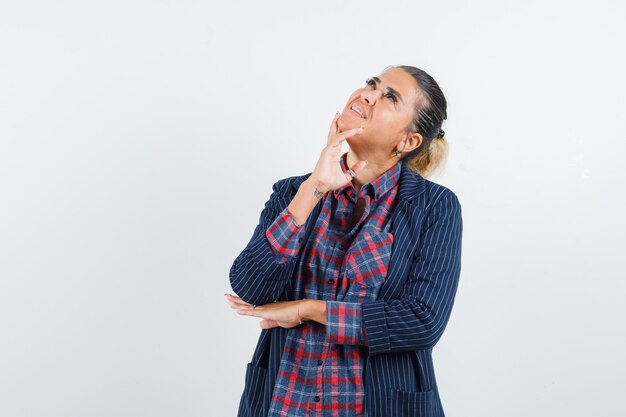 Blonde lady in shirt, jacket holding fingers on chin and looking dreamy , front view.