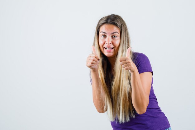 Блондинка показывает двойные пальцы вверх в фиолетовой футболке и выглядит радостной, вид спереди.