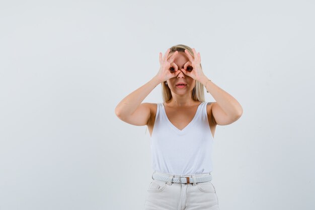 Blonde lady showing glasses gesture in singlet, pants and looking focused