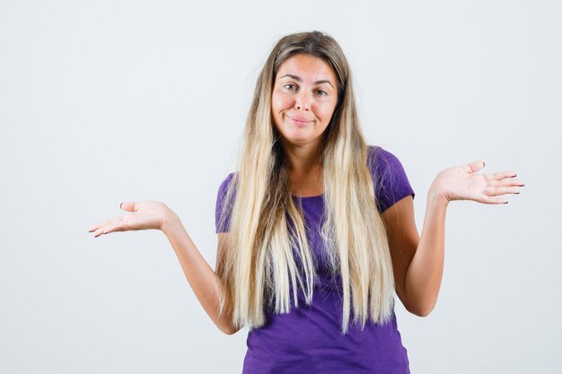 Blonde lady showing helpless gesture in violet t-shirt and looking confused. front view.