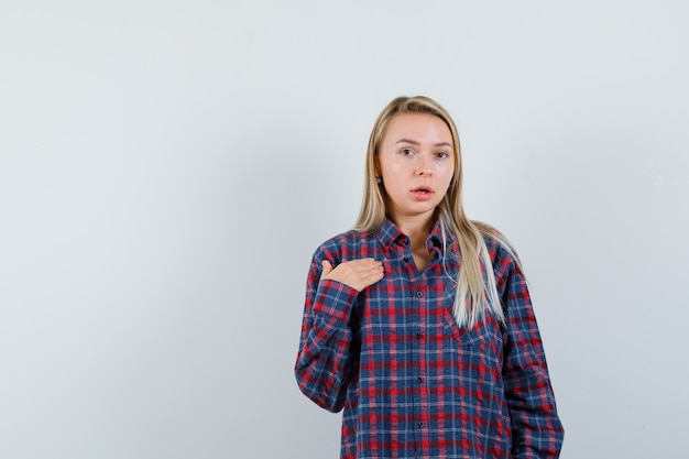 Signora bionda che mostra se stessa in modo interrogativo in camicia casual e guardando perplessa, vista frontale.