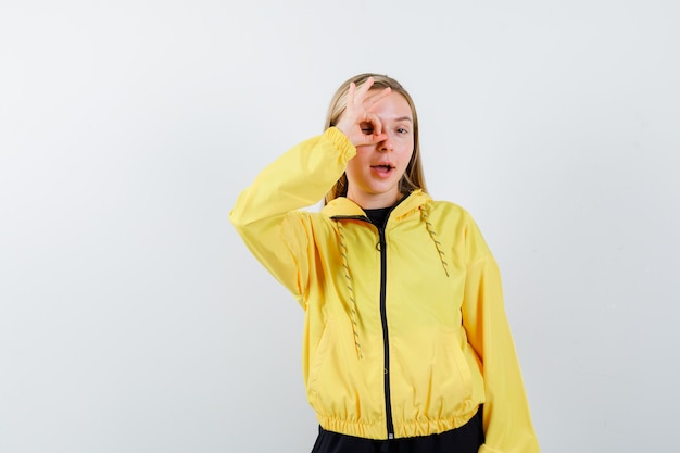 Blonde lady showing ok sign on eye in tracksuit and looking amazed , front view.