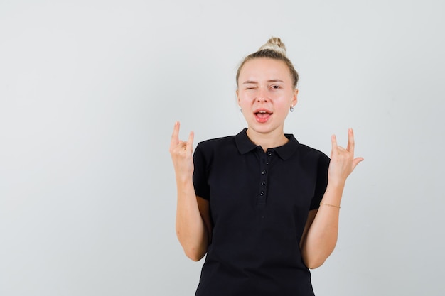 Blonde lady showing rock n roll sign, winking eye in black t-shirt and looking happy. front view.