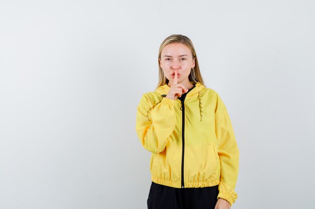Blonde lady showing silence gesture in tracksuit and looking sensible , front view.