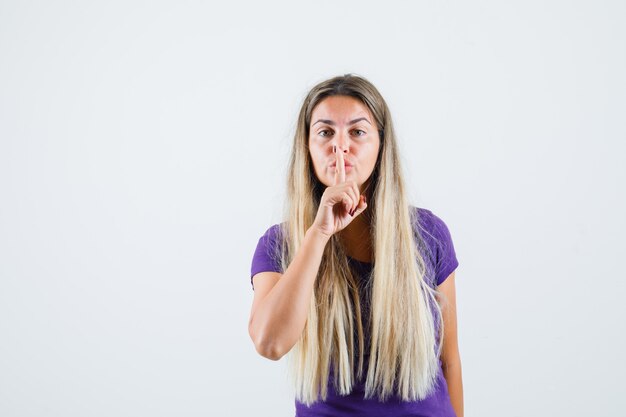 Blonde lady showing silence gesture in violet t-shirt and looking careful , front view.