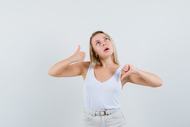 Blonde lady showing thumbs up an down in singlet, pants and looking indecisive , front view.