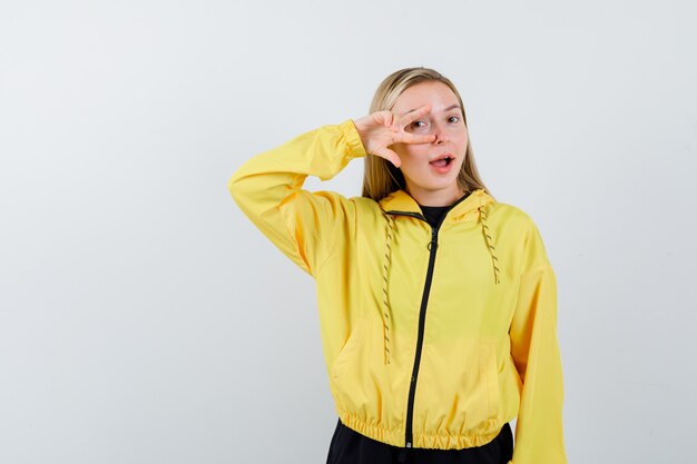 Blonde lady showing V-sign on eye in tracksuit and looking amazed. front view.