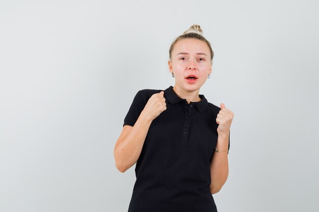 Blonde lady showing winner gesture in black t-shirt and looking happy. front view.