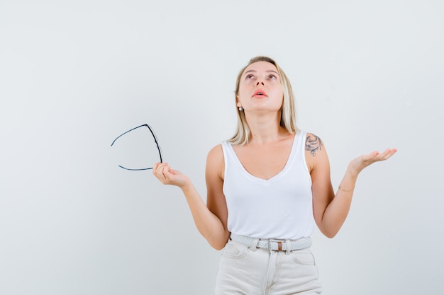 Blonde lady in singlet, pants holding glasses, looking up and looking hopeful , front view.