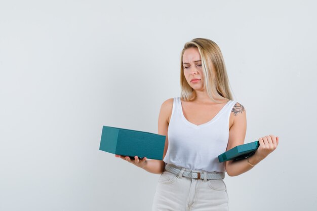 Blonde lady in singlet, pants looking into present box and looking disappointed , front view.