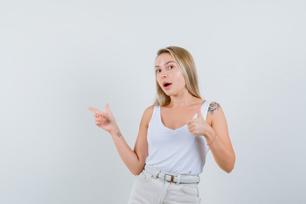 Blonde lady in singlet, pants pointing aside, showing thumb up and looking optimistic, front view.