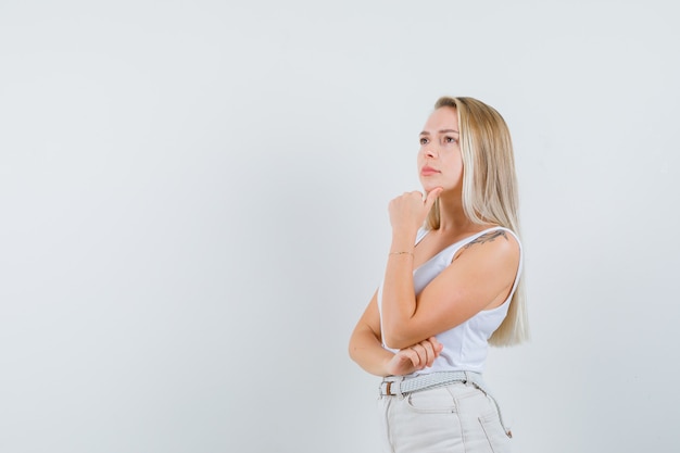 Blonde lady in singlet, pants propping chin on hand and looking thoughtful .