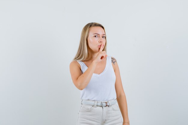 Blonde lady in singlet, pants showing silence gesture and looking careful , front view.