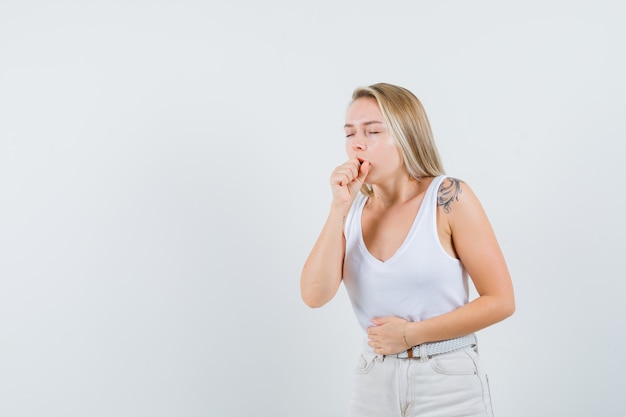 Blonde lady in singlet, pants suffering from cough and looking sick , front view.