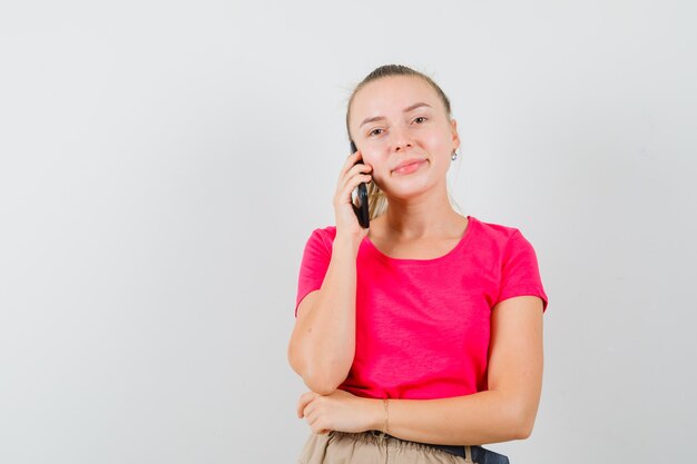 Blonde lady smiling while talking on mobile phone in pink t-shirt front view.