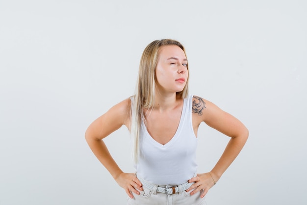 Blonde lady standing to hear clearly in singlet, pants and looking curious , front view.