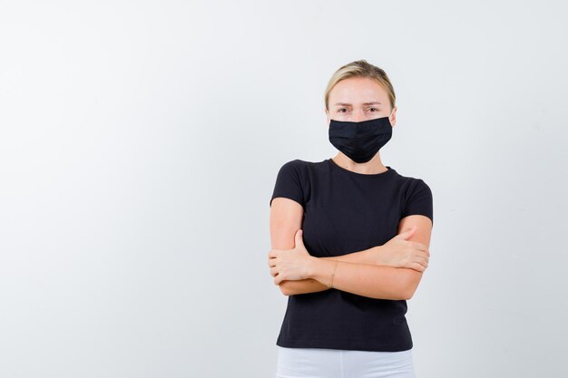 Blonde lady standing with crossed arms in black t-shirt, black mask isolated