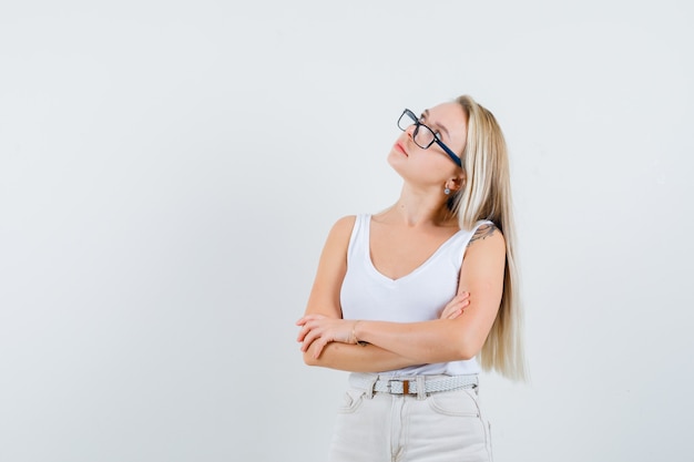 Blonde lady standing with crossed arms in singlet, pants and looking pensive. front view.