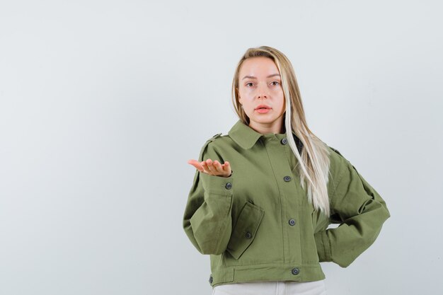 Бесплатное фото Блондинка протягивает руку озадаченным жестом в куртке, штанах, вид спереди.