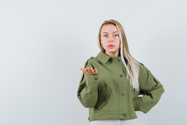 Blonde lady stretching hand in puzzled gesture in jacket, pants , front view.