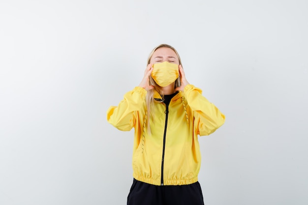 Blonde lady suffering from headache in tracksuit, mask and looking annoyed. front view.