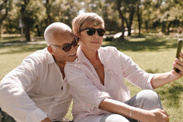 Blonde lady in sunglasses, striped stylish blouse and jeans. sitting on grass and making selfie with mustachioed man in white shirt outdoor.