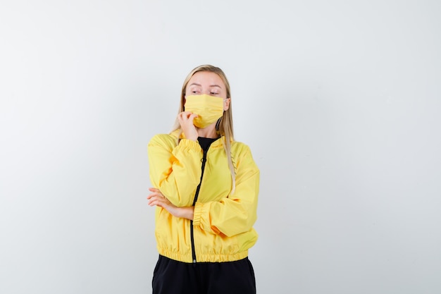 Blonde lady in tracksuit, mask holding hand on chin and looking pensive , front view.