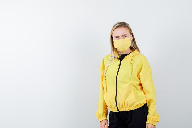 Blonde lady in tracksuit, mask looking at camera and looking nervous , front view.