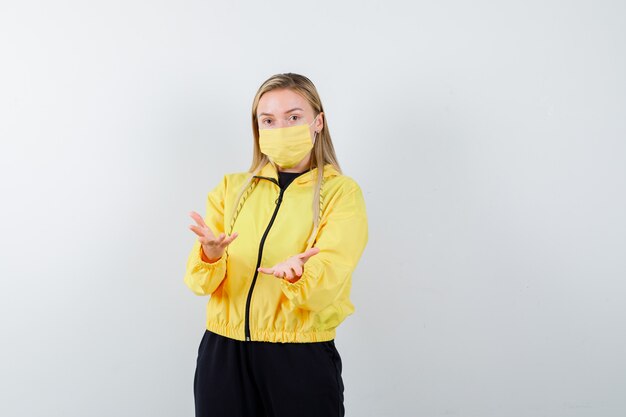 Blonde lady in tracksuit, mask pretending to present something and looking puzzled , front view.