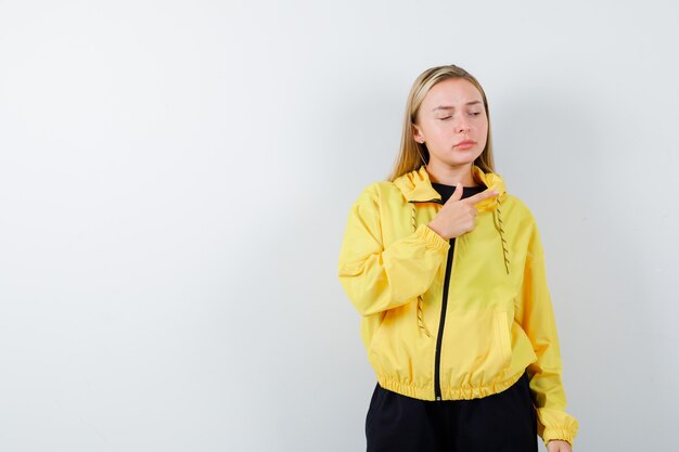Blonde lady in tracksuit pointing right and looking sad , front view.