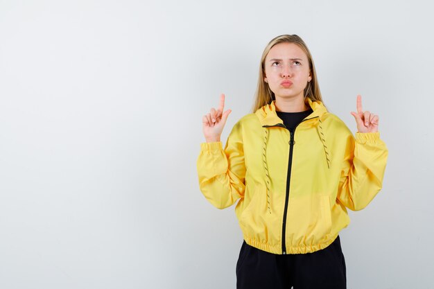 Blonde lady in tracksuit pointing up, keeping lips folded and looking hesitant , front view.