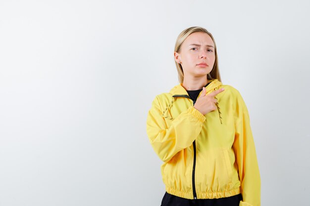 Blonde lady in tracksuit pointing at upper right corner and looking hesitant , front view.