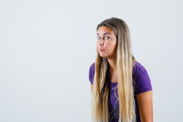 Blonde lady in violet t-shirt overhearing private conversation and looking curious , front view.