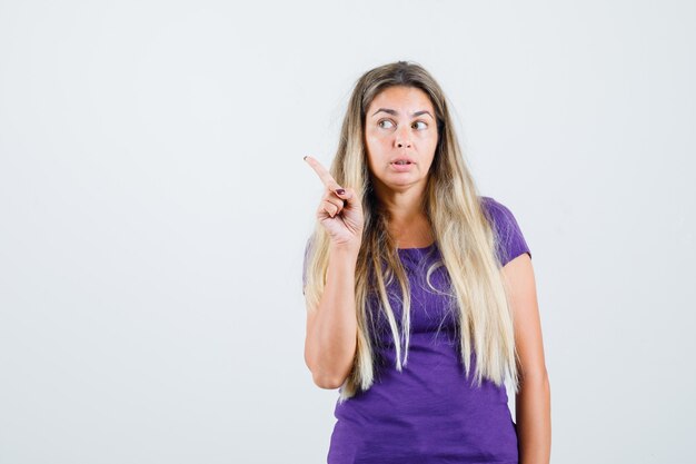 Blonde lady in violet t-shirt pointing away and looking curious , front view.