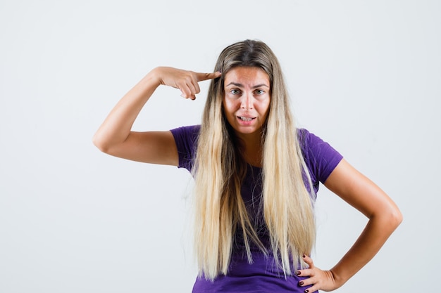 Blonde lady in violet t-shirt pointing at her temples , front view.