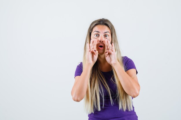 Blonde lady in violet t-shirt telling secret with hands near mouth , front view.