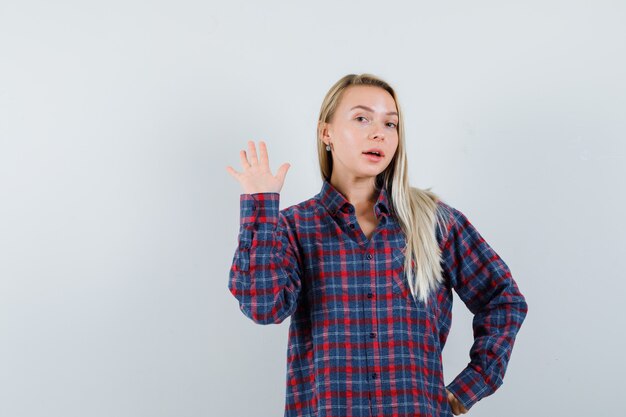 Blonde lady waving hand to say goodbye in casual shirt and looking confident. front view.