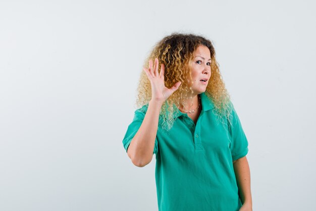 Blonde lady with curly hair in green T-shirt overhearing private conversation and looking curious , front view.