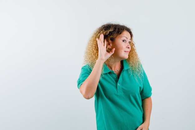 Blonde lady with curly hair keeping hand behind ear, overhearing secret in green T-shirt and looking curious , front view.