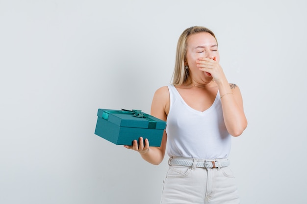 Blonde lady yawning, holding present box in singlet, pants and looking sleepy. front view.