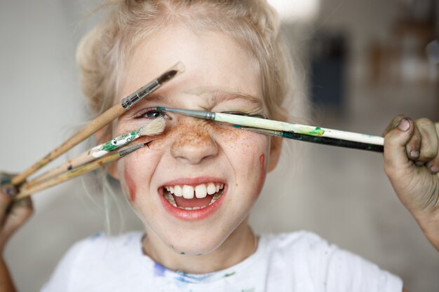 Blonde little girl in playful mood with paint on her freckled face and blue eyes covering her face with brushes