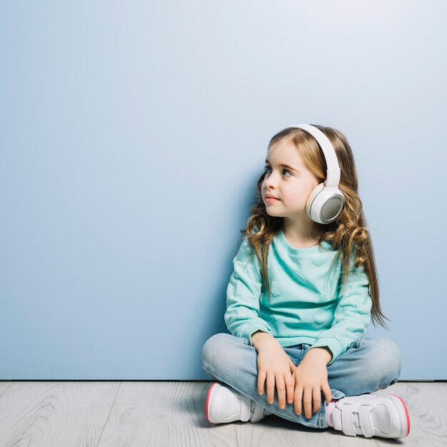 Blonde little girl sitting on hardwood floor listening music on headphone looking away