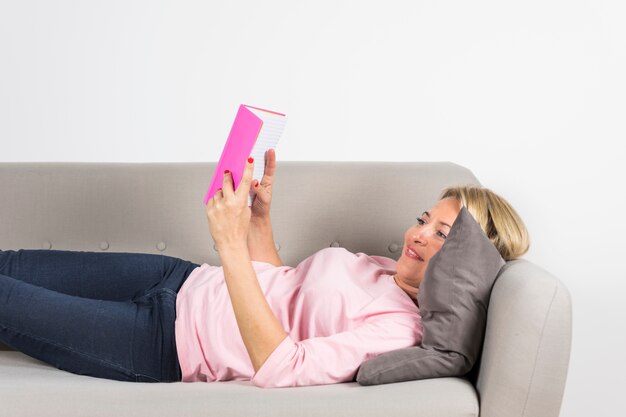 Blonde mature woman lying on sofa reading book against white backdrop