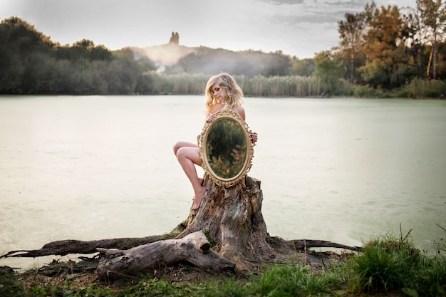 Blonde naked woman holds a mirror sitting before a lake covered with mist