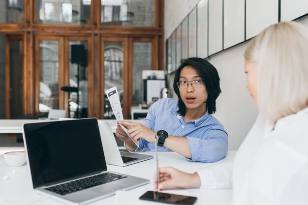 금발 비서 검은 화면 노트북 옆에 앉아 안경에 아시아 젊은 남자를 듣고. 갈색 머리 중국 회사원 흰 블라우스에 여성 관리자와 이야기.