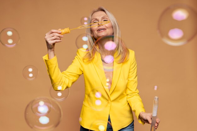 Blonde senior woman blowing bubbles against a yellow background