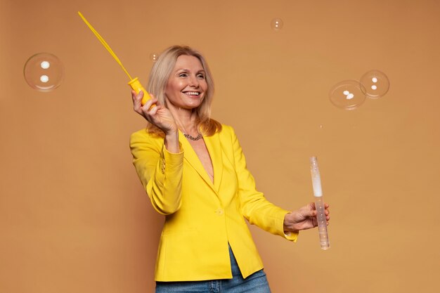 Blonde senior woman blowing bubbles and smiling on a yellow background