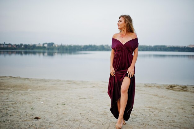 Blonde sensual barefoot woman in red marsala dress posing against lake on sand