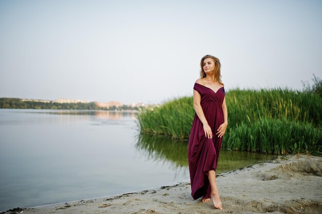 갈대와 호수에 대 한 포즈를 취하는 빨간 마르살라 드레스에 금발의 관능적인 맨발의 여자